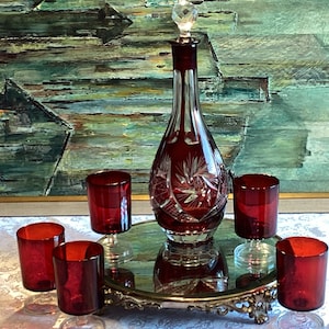 Puede incluir: Un juego de cinco vasos de cristal rojo, incluyendo una jarra con un tapón de cristal transparente, sobre una bandeja de metal dorado con una superficie de espejo.