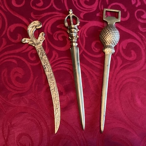 May include: Three antique letter openers in a row. The openers are made of gold-colored metal. The first is curved with an ornate handle. The second has a detailed handle. The third has a pineapple-shaped handle.