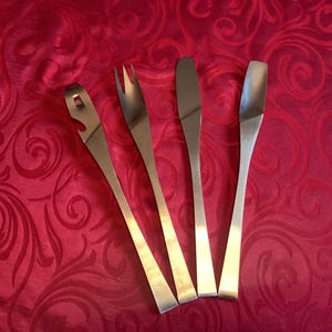 May include: A set of four stainless steel serving utensils, including a bottle opener, fork, knife, and spoon. The utensils are arranged on a red patterned tablecloth.