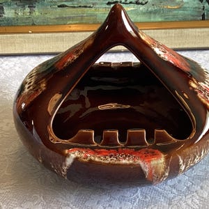 May include: A brown ceramic ashtray with a unique design. The ashtray has a triangular opening in the centre and a raised rim around the edge. The ashtray is decorated with a brown, white and red glaze.