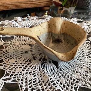 Puede incluir: Un cucharón de cerámica con un acabado moteado en beige y gris. La pieza presenta un mango largo y plano con un orificio circular en el extremo y un cuenco profundo y redondeado con un pico vertedor distintivo en el lateral, de estilo rústico.