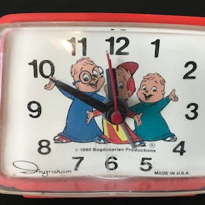 Pode incluir: Um despertador retangular vermelho com um mostrador branco com os personagens de desenho animado Alvin, Simon e Theodore. O relógio tem números e ponteiros pretos. O relógio está marcado "Made in U.S.A."