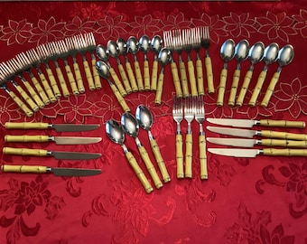 May include: A collection of stainless steel silverware with bamboo-style handles. The set includes forks, spoons, and knives, arranged on a red tablecloth with a floral pattern. The bamboo handles have a light yellow color.