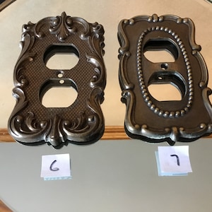 May include: Two ornate metal outlet covers in a bronze finish. The cover on the left has a more intricate design with scrollwork and floral motifs. The cover on the right has a simpler design with a beaded border. Both covers are labeled with numbers, 6 and 7.