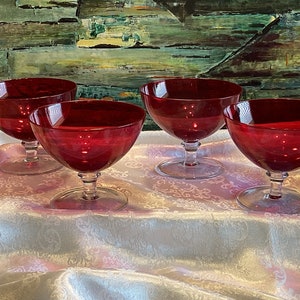 May include: Four red glass dessert dishes with clear stems and bases. The dishes are arranged on a white tablecloth with a floral pattern.