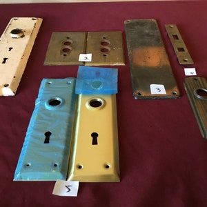 May include: Collection of vintage door hardware, including various plates and escutcheons. The pieces are in different colors and finishes, such as white, brass, and aged metal. Some are new, while others show signs of wear.