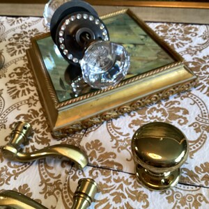 May include: A collection of door hardware including a clear faceted glass knob with a dark base ringed by small clear stones, a polished brass round knob, and two brass lever handles, all arranged near a rectangular gold-framed mirror base.