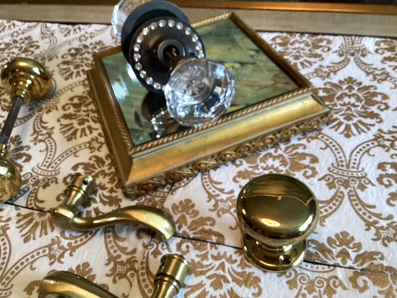 May include: A collection of door hardware including a clear faceted glass knob with a dark base ringed by small clear stones, a polished brass round knob, and two brass lever handles, all arranged near a rectangular gold-framed mirror base.