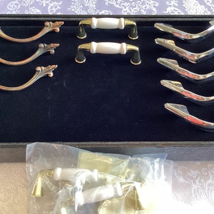 Puede incluir: Surtido de herrajes para armarios en una vitrina de terciopelo negro. El herraje incluye tres juegos de tiradores de cajones ornamentados de color cobre, dos juegos de tiradores de cajones de cerámica blanca con detalles dorados y cinco juegos de tiradores de cajones cromados.