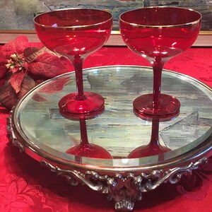Peut inclure: Deux coupes à champagne en verre rouge avec des bords dorés sont posées sur un plateau de service en argent miroir avec des détails ornés. Le plateau a un dessus rond et un bord festonné.