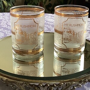 May include: Two clear glass tumblers with gold trim and a frosted white surface. The tumblers have a gold inscription that reads 
