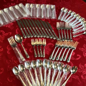 Puede incluir: Una colección de cubiertos plateados, que incluye tenedores, cuchillos y cucharas, dispuestos sobre un mantel rojo floral. Algunas piezas están envueltas individualmente en plástico. La cubertería tiene un diseño decorativo en el mango.