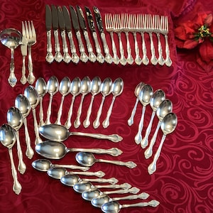 May include: A set of silver flatware, including knives, forks, spoons, and serving utensils, arranged on a red tablecloth. The set includes a variety of sizes and styles of spoons, including soup spoons, teaspoons, and tablespoons.
