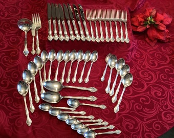 May include: A set of silver flatware, including knives, forks, spoons, and serving utensils, arranged on a red tablecloth. The set includes a variety of sizes and styles of spoons, including soup spoons, teaspoons, and tablespoons.