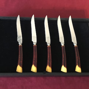 May include: Set of six stainless steel steak knives with brown and yellow handles. The knives are displayed in a black velvet-lined box.