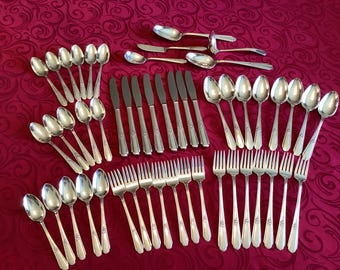 May include: A collection of silver-colored flatware, including spoons, forks, and knives, arranged on a red patterned tablecloth. The silverware has a decorative design on the handles. The set includes serving spoons.