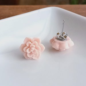 May include: A pair of light pink flower-shaped stud earrings. The earrings are made of a smooth material and have a detailed petal design. The earring posts are silver-colored. The earrings are displayed on a white surface.