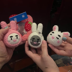 Puede incluir: Tres llaveros de conejito de ganchillo hechos a mano en rosa, blanco y blanco con caras grises. Un marco de fotos con forma de fresa con una tarjeta azul está en el fondo. Los llaveros están sostenidos por manos.