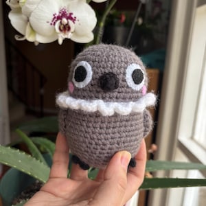 Könnte beinhalten: Eine gehäkelte graue Eule mit weißen Akzenten um den Hals und schwarzen Augen. Die Eule wird in einer Hand gehalten.