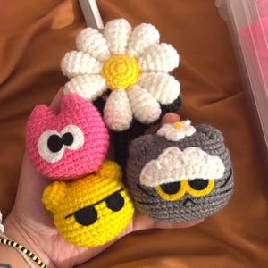 May include: A selection of handmade crochet items. The image shows a large white daisy with a yellow centre, and several small, colourful character-shaped pieces. These include a pink creature, a yellow bear with sunglasses, and a grey cat.
