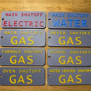 May include: Eight rectangular gray plastic tags with holes for mounting. The tags feature raised lettering: MAIN SHUTOFF ELECTRIC in red, MAIN SHUTOFF WATER in blue, and six tags labeled MAIN SHUTOFF GAS, DRYER SHUTOFF GAS, FURNACE SHUTOFF GAS, STOVE SHUTOFF GAS, OVEN SHUTOFF GAS, and WATER HEATER SHUTOFF GAS in yellow.
