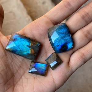 Pode incluir: Quatro cabochões de labradorita, pedras preciosas iridescentes, de diferentes tamanhos e formas, com um brilho azul-esverdeado e dourado. As pedras são exibidas em uma mão.