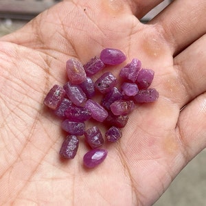 May include: A collection of small, rough, pink ruby gemstones. The rubies are various shades of pink and have a natural, unpolished look.