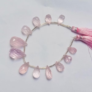 May include: A strand of pink rose quartz gemstone beads, each bead is faceted and shaped like a teardrop. The beads are strung on a silver wire and have a pink tassel on each end.