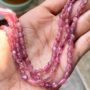 May include: A strand of pink gemstone beads, likely tourmaline, with a faceted, elongated shape. The beads are strung on a thin cord and are displayed on a hand.