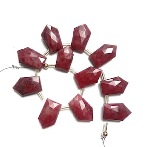 Puede incluir: Una hebra de cuentas de piedras preciosas rojas con forma facetada, alargada y rectangular. Las cuentas están enhebradas en un alambre de plata con separadores de color dorado.