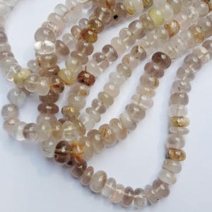 May include: Strands of gemstone beads in varying shades of brown, tan, and clear. The beads are small, rounded, and strung together, creating a necklace or bracelet. The beads have a natural, organic appearance, with a slightly translucent quality.