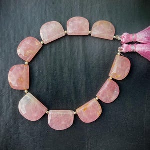 May include: A strand of pink, semi-precious gemstone beads. The beads are a soft pink color, cut into a half-moon shape, and strung together with small silver-colored beads. A pink tassel is attached to the end of the strand.
