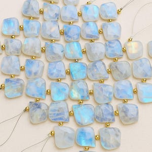 May include: Close-up of a strand of moonstone beads. Each bead is a square shape with a faceted surface, displaying a mix of white and blue hues. The beads are strung together with gold-colored spacers and thin, clear string.