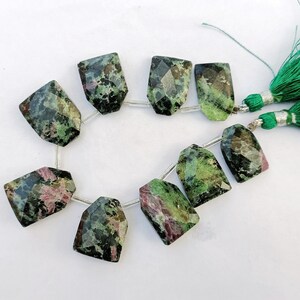 Puede incluir: Un hilo de cuentas de turmalina facetadas y rectangulares en tonos verdes, negros y rosas. Las cuentas están ensartadas en un alambre plateado, con una borla verde en el lado derecho.