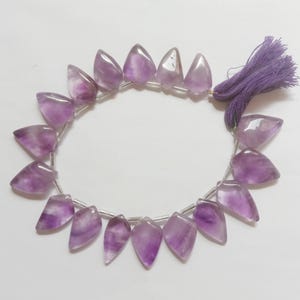 May include: A strand of purple amethyst gemstone beads, each bead is faceted and shaped like a teardrop. The beads are strung on a silver wire and have a purple tassel at the end.