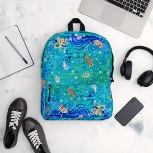 Könnte beinhalten: Ein türkisfarbener Rucksack mit einem Meeresleben-Muster mit Oktopussen, Quallen und anderen Meereskreaturen. Der Rucksack hat einen schwarzen Griff und Reißverschluss und ist von einem Laptop, Kopfhörern, einem Telefon, einem Notizbuch und schwarzen Turnschuhen umgeben.