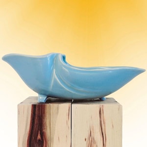 May include: A light blue ceramic planter with a smooth, glossy finish and an elongated, curved boat-like shape. The vessel features a wide, open top and is supported by small, integrated feet, showcasing a fluid, sculptural design throughout.