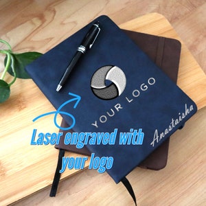 Könnte beinhalten: Ein blaues, ledergebundenes Notizbuch mit einem schwarzen Stift darauf. Das Notizbuch hat einen silbernen Kreis mit einem Design darin und den Text "YOUR LOGO" darunter. Der Text "Laser engraved with your logo" ist in Blau auf weißem Hintergrund geschrieben. Das Notizbuch liegt auf einem braunen, ledergebundenen Notizbuch.