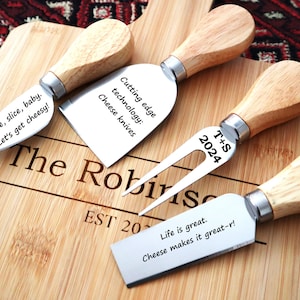 May include: A bamboo serving board engraved with The Robinson EST 2024, featuring three stainless steel cheese knives with light wood handles. The blades are engraved with the phrases Slice, slice, baby. Let's get cheesy!, Cutting edge technology: Cheese knives, T+S 2024, and Life is great. Cheese makes it great-r!.