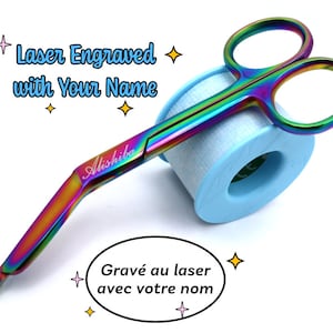 Pode incluir: Tesoura cirúrgica arco-íris com cabo curvo. A tesoura está apoiada em um rolo de fita médica azul. A tesoura é gravada com o nome "Alishiba" e o texto "Gravado a laser com seu nome".