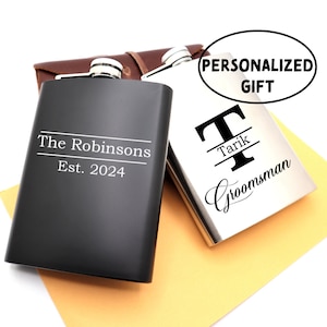May include: Two personalised stainless steel hip flasks. One flask is black with the text "The Robinsons Est. 2024". The other flask is silver with the text "T Tarik Groomman".