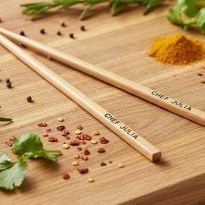 Peut inclure: Deux baguettes en bois avec l'inscription "CHEF JULIA", posées sur une planche à découper en bois. La planche est parsemée d'herbes, d'épices et de grains de poivre, évoquant la cuisine.