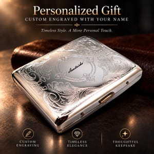 May include: A rectangular silver-toned metal cigarette case featuring an intricate embossed floral scrollwork pattern across the surface. The center of the case displays the name "Anastaisha" engraved in a cursive script within an oval frame.