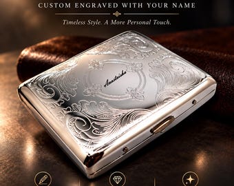 May include: A rectangular silver-toned metal cigarette case featuring an intricate embossed floral scrollwork pattern across the surface. The center of the case displays the name "Anastaisha" engraved in a cursive script within an oval frame.