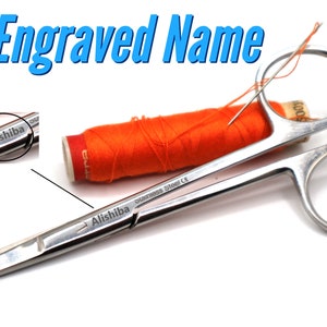 May include: A pair of silver surgical scissors with the name "Alishiba" engraved on the handle. The scissors are lying on a white surface next to an orange spool of thread and a needle.