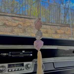 May include: A car interior with a rearview mirror hanging crystal charm. The charm is made of three pink, white, and purple crystals tied together with string and a cream-colored tassel.