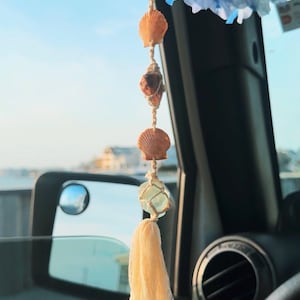May include: A car charm featuring seashells, crystals, and a cream-colored tassel. The charm is suspended from a string and hangs in front of a car window. Blue and pink flowers are visible at the top.