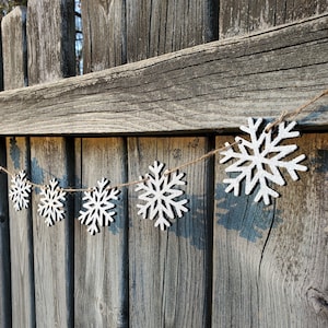 May include: A white snowflake garland with a rustic twine string hanging on a weathered wooden fence. The snowflakes are made of wood and have a slightly distressed look.