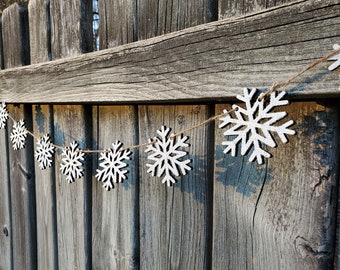 May include: A white snowflake garland with a rustic twine string hanging on a weathered wooden fence. The snowflakes are made of wood and have a slightly distressed look.