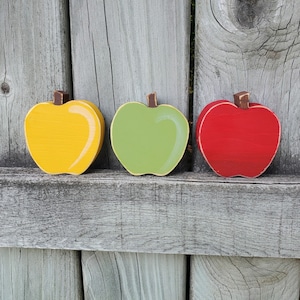 May include: Three wooden apples painted in red, green, and yellow. The apples are arranged in a row on a wooden surface.